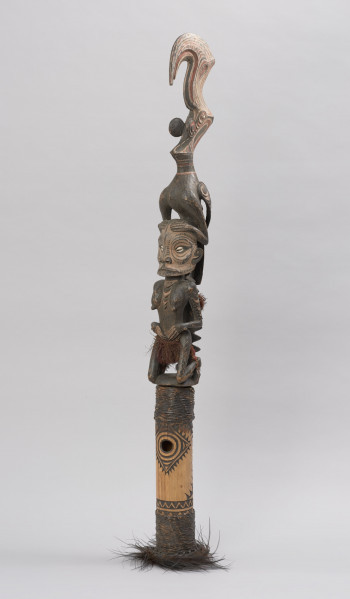 flet obrzędowy - Ujęcie ze skosu z lewej; Podłużny, bogato zdobiony flet wykonany z odcinka bambusa otwartego po stronie służącej do zadęcia i zamkniętego naturalnym kolankiem po stronie przeciwnej, gdzie znajduje się drewniana figura kobiety-przodka w pozycji klęczącej z ptakiem na głowie.