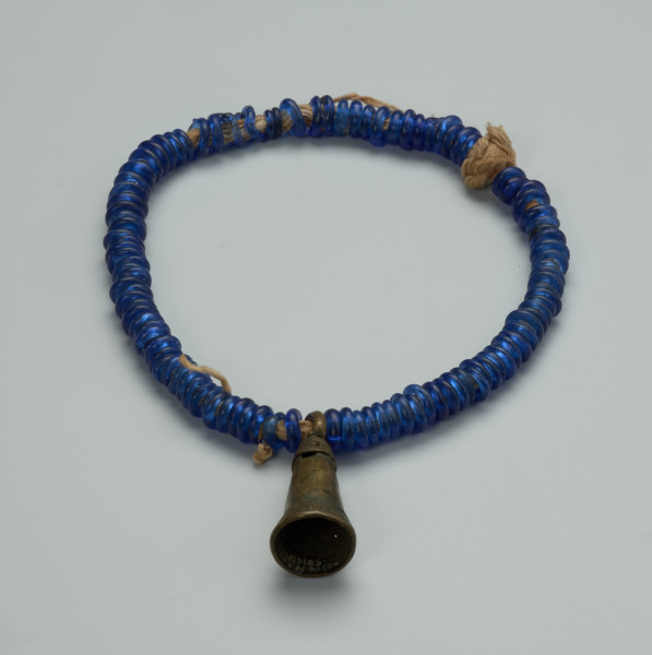 biżuteria, naszyjnik - Ujęcie z tyłu. Zamknięty, kobaltowy naszyjnik. Na bawełnianym sznurku znajdują się nałożone szklane, kobaltowe paciorki oraz wykonany z brązu wisiorek w kształcie wydłużonego stożka przypominającego dzwon. Wisiorek ozdobiono wzorami geometrycznymi.