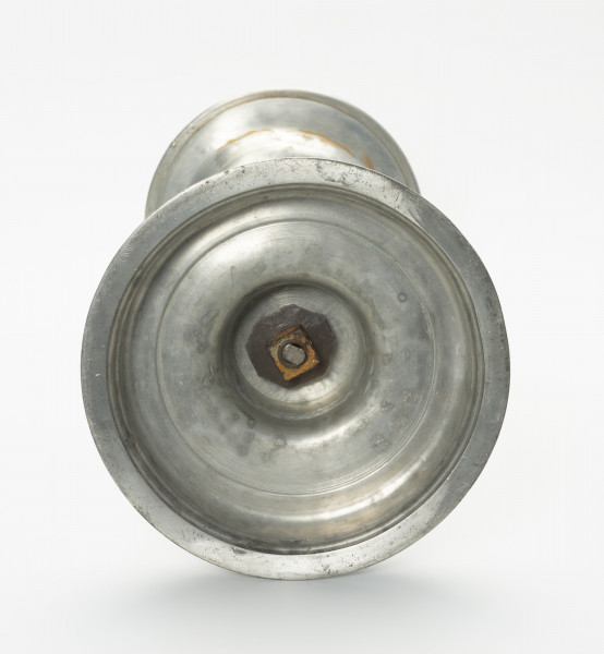 cynowy świecznik na okrągłej, wysklepionej stopie - Ujęcie od spodu; Cynowy świecznik na okrągłej, wysklepionej stopie. Spód świecznika z mocowaniem od spodu. Słabo widoczne tłoczone zdobienia - pieczęcie.