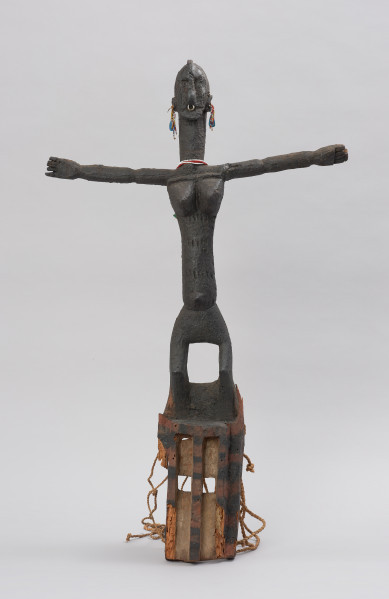 rzeźbiona maska - Ujęcie z przodu. Drewniana maska z dużymi otworami na oczy, do której przymocowany jest bawełniany sznurek. Maska przyozdobiona jest wyrzeźbioną postacią kobiety z podniesionymi rękami.