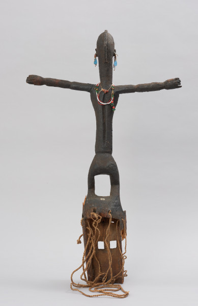 rzeźbiona maska - Ujęcie z tyłu. Drewniana maska z dużymi otworami na oczy, do której przymocowany jest bawełniany sznurek. Maska przyozdobiona jest wyrzeźbioną postacią kobiety z podniesionymi rękami.