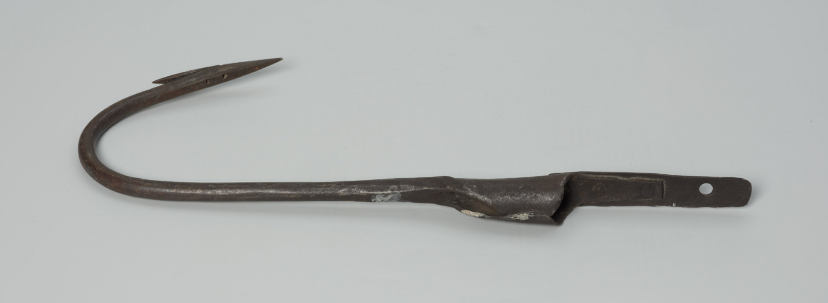 harpun - Ujęcie tyłu w poziomie. Duży, wykonany z żelaza techniką kowalską harpun. Część pracująca łukowato wygięta, zaostrzona z przygwintowanym pojedynczym, ruchomym  zadziorem. Zakończona tuleją, w której osadzona była rękojeść (rękojeści brak).  Z tulei wyprowadzony krótki pas metalowy, do którego za pomocą trzech nitów, po zewnętrznej stronie, mocowany jest pas dłuższy. W tulei i w dłuższym pasie wycięte, znajdujące się na jednej linii, okrągłe otwory.