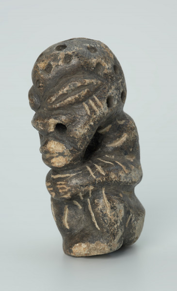 rzeźba; przedmiot obrzędowy; Figura kultu zmarłych - Ujęcie z przodu z lewej strony. Rzeźbiona w grafitowobeżowym steatycie siedząca postać ludzka, której nieproporcjonalnie dużą głowę zdobią liczne, okrągłe wgłębienia, natomiast ciało pokrywają pionowe i ukośne nacięcia, oznaczające prawdopodobnie skaryfikację. Powierzchnia rzeźby z rysami, mikropęknięciami, przetarciami oraz drobnymi uszkodzeniami.