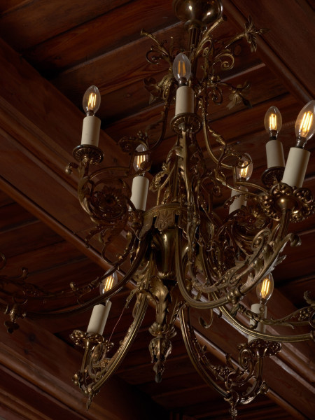 żyrandol - Ujęcie z lewej strony, zbliżenie. Żyrandol 12-ramienny, mosiężny z kryształowymi soplami.Trzon w górnej części o przekroju kwadratowym, ozdobiony wypukłą dekoracją o formach geometrycznych i roślinnych, w dolnej części tralkowy z zakończeniem w kształcie szyszki. Ramiona w formie gałązek wygiętych półkoliście, połączonych przy trzonie w 4 wiązki po 3 elementy: dwa dłuższe po bokach i krótsze w środku. Każda trójka przechodzi przy trzonie w jedną spiralnie zwiniętą gałązkę akantu. Środkowe części ramion ozdobione są analogicznymi motywami: łodyżką z listkami rozszczepiającą się na dwa spiralnie skręcone pędy. Profitki ażurowe, zbudowane z ułożonych w rozetkę listków akandu. Przy górnej części trzonu, w połowie wysokości motyw dekoracyjny złożony z dwóch wygiętych, a na końcach spiralnie skręconych gałązek akantu. Górny talerz maskujący pod sufitem zawieszkę - płaski w formie kwiatu akantu. Ramiona i trzon połączone luźno zwieszonymi łańcuszkami, umieszczonymi także przy talerzu podsufitowym i ozdobach górnej części trzonu. Przy profitkach szklane wisiorki: 16 sztuk szlifowanych w kształcie sopelków - podłużne o trójkątnym przekroju z ostrosłupowym zakończeniem oraz 5 sztuk w kształcie pękatych łezek. Świece tulejowe z tworzywa sztucznego, zaopatrzone we wkręty do żarówek świecowych. Wewnętrzna instalacja elektryczna.