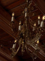 żyrandol - Ujęcie z lewej strony, zbliżenie. Żyrandol 12-ramienny, mosiężny z kryształowymi soplami.Trzon w górnej części o przekroju kwadratowym, ozdobiony wypukłą dekoracją o formach geometrycznych i roślinnych, w dolnej części tralkowy z zakończeniem w kształcie szyszki. Ramiona w formie gałązek wygiętych półkoliście, połączonych przy trzonie w 4 wiązki po 3 elementy: dwa dłuższe po bokach i krótsze w środku. Każda trójka przechodzi przy trzonie w jedną spiralnie zwiniętą gałązkę akantu. Środkowe części ramion ozdobione są analogicznymi motywami: łodyżką z listkami rozszczepiającą się na dwa spiralnie skręcone pędy. Profitki ażurowe, zbudowane z ułożonych w rozetkę listków akandu. Przy górnej części trzonu, w połowie wysokości motyw dekoracyjny złożony z dwóch wygiętych, a na końcach spiralnie skręconych gałązek akantu. Górny talerz maskujący pod sufitem zawieszkę - płaski w formie kwiatu akantu. Ramiona i trzon połączone luźno zwieszonymi łańcuszkami, umieszczonymi także przy talerzu podsufitowym i ozdobach górnej części trzonu. Przy profitkach szklane wisiorki: 16 sztuk szlifowanych w kształcie sopelków - podłużne o trójkątnym przekroju z ostrosłupowym zakończeniem oraz 5 sztuk w kształcie pękatych łezek. Świece tulejowe z tworzywa sztucznego, zaopatrzone we wkręty do żarówek świecowych. Wewnętrzna instalacja elektryczna.