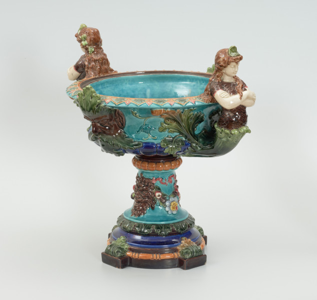 Waza - Ujęcie z przodu skosem w prawą stronę. Waza w kształcie smukłej amfory. Brzusiec ceramiczny, rozszerzający się ku górze, w kolorze kremowym, malowany podszkliwnie (z uzupełnieniami naszkliwnymi) we wzory kwiatowe - bukiety barwnych kwiatów w typie aster w kolorach fioletowo-czerwonym, liliowym, żółtym i białym, z gałązkami i listkami oraz drobnymi kwiatkami przypominającymi bratki o barwie fioletowo-czerwonej i liliowej. Górna część wazy wraz z uchwytami oraz stopą i część podstawy wykonane jako odlewy z mosiądzu. Mosiężna szyja wąska z otokiem perełkowym w środku ozdobiona jest pionowym, rozszerzającym się ku dołowi i ku górze rytmicznym podziałem z ornamentem geometryczno-roślinnym. Na pogrubionym skraju szyi umieszczona jest płytka miseczka w formie kwiatu o wąskich płatkach, stanowiąca wylewkę wazonu. Poniżej dolnej części szyi oprawa wierzchołka brzuśca składająca się z otoku o sercowym wycięciu zwieńczonym profilowaną palmetką oraz dwóch wydłużonych, sięgających 1/3 wysokości brzuśca, pionowych wsporników uchwytów. Te ostatnie składają się z 2 części: zewnętrzna wychodząca ze wsporników opartych na brzuścu, wyniesiona jest po niewielkim skosie do połowy wysokości szyi i na końcu wywinięta do wnętrza wolutowo, a druga - krótsza, skierowana ku dołowi łączy łagodnym łukiem wolutę z górną częścią oprawy brzuśca, kończąc się wywiniętymi na zewnątrz listkami. Uchwyty żłobkowane, zdobione motywem liści. Wazon wsparty jest na mosiężnej podstawie, składającej się z wieńca oplecionego wstęgą, na którym spoczywa zwężająca się ku górze stopa o brzegach ozdobionych perełkowaniem i pierścieniem, wspierającym koszyczek, ujmujący dolną część ceramicznego brzuśca, w formie kwiatu o ostro zakończonych, wąskich płatkach. Całość spoczywa na podstawie z kwadratowej płytki czerwonego marmuru ze ściętymi narożnikami, ujętymi mosiężnymi wspornikami ramy wspierającej płytę od dołu i opartej w narożach na czterech lwich łapach.