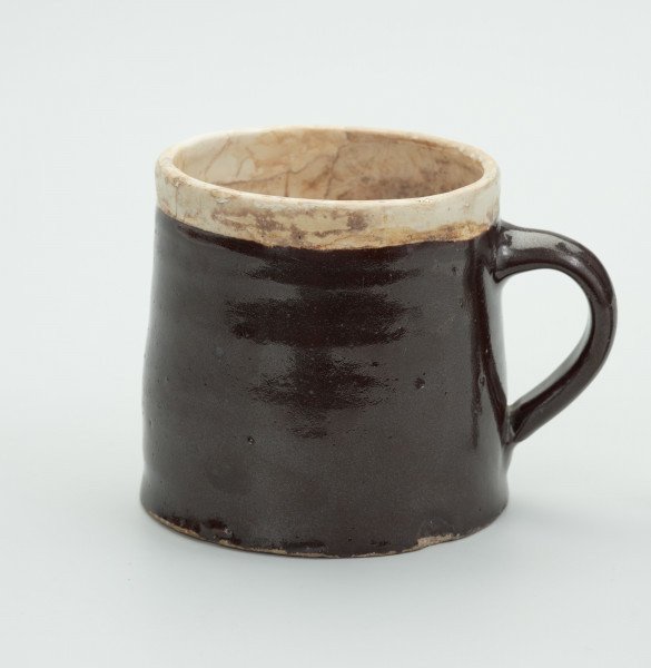 Kamionkowy kubek w kształcie kufla - Ujęcie z lewej strony; Kubek w kształcie walca zwężającego się ku górze. Wlew węższy od dna. Szkliwo dwukolorowe; wokół wylewu i w środku jasnobeżowe/kremowe, pozostała część i ucho w kolorze ciemnego brązu. Ucho wzdłużne, taśmowe umieszczone od linii jasnego szkliwa i zakończone na 4/5 wysokości naczynia.