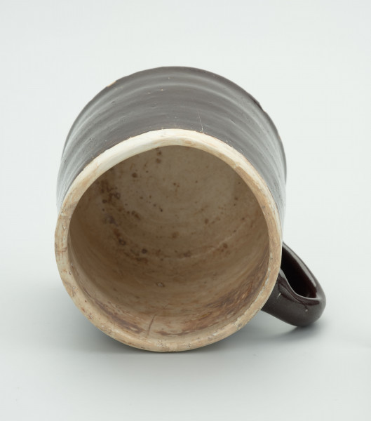 Kamionkowy kubek w kształcie kufla - Ujęcie wnętrza; Kubek w kształcie walca zwężającego się ku górze. Wlew węższy od dna. Szkliwo dwukolorowe; wokół wylewu i w środku jasnobeżowe/kremowe, pozostała część i ucho w kolorze ciemnego brązu. Ucho wzdłużne.