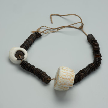 biżuteria, naszyjnik - Ujęcie z przodu. Naszyjnik typu otwartego, na który nałożone są drewniane paciorki w kolorze brązowym o płaskim i okrągłym kształcie z małym otworem pośrodku. Całość przedzielono dużą i mniejszą muszlą kauri, które pełnią rolę wisiorków.