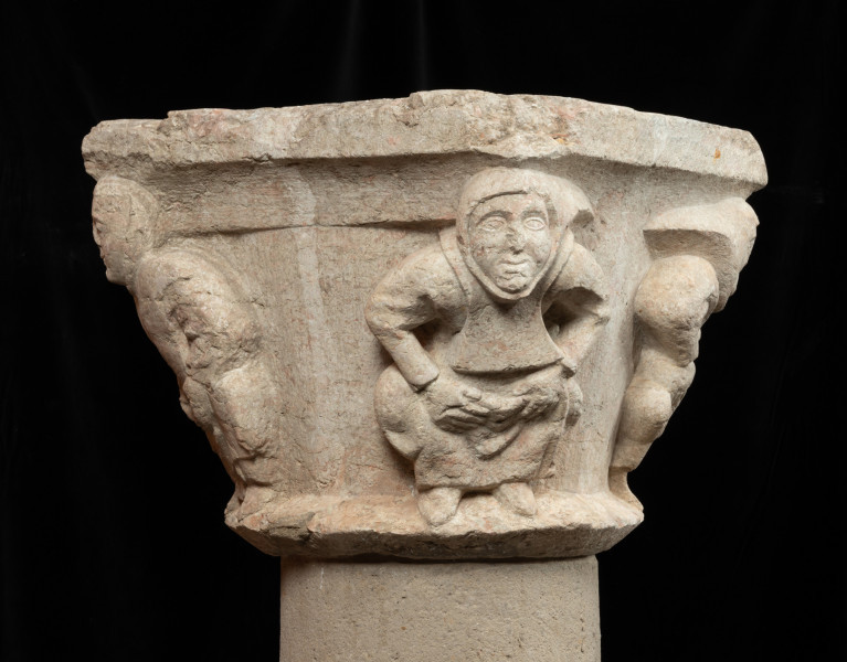 Kapitel z przedstawieniem czterech mnichów - atlantów - Ujęcie drugiego boku. Kapitel blokowo-kielichowy, rozszerzający się ku górze, wykuty z jednego kawałka kamienia. Okrągłą podstawę otacza wałek o trójkątnym profilu. Zwieńczenie w formie szerokiej czworobocznej płyty abakusa. W narożnikach kapitela płaskorzeźbione wizerunki czterech mnichów. Postaci o krępej budowie ukazane frontalnie, w pozycji przykucniętej. Stopami opierają się o dolny wałek, głowami podpierają abakus. Relief głęboki, półplastyczny, światłocieniowy. Gładko opracowane tło. Niewielkie uszczerbienia zwłaszcza w partii abakusa i dolnego wałka. Pod spodem centralne wgłębienie do umocowania trzpienia łączącego kapitel z trzonem.