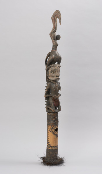 flet obrzędowy - Ujęcie ze skosu z prawej; Podłużny, bogato zdobiony flet wykonany z odcinka bambusa otwartego po stronie służącej do zadęcia i zamkniętego naturalnym kolankiem po stronie przeciwnej, gdzie znajduje się drewniana figura kobiety-przodka w pozycji klęczącej z ptakiem na głowie.