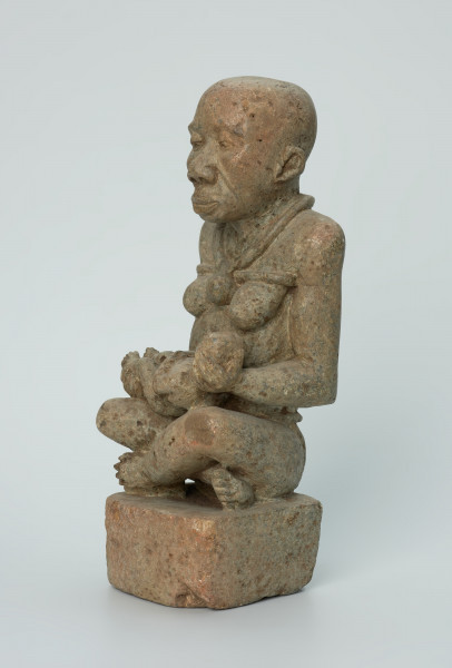 Ujęcie z przodu z lewej. Rzeźbiona w biało-szarym kamieniu dość duża postać kobiety w pozycji siedzącej z nogami skrzyżowanymi. Kobieta siedzi na małej, kwadratowej, lekko zaokrąglonej na rogach podstawie, w dłoniach trzyma dziecko. Widoczne rysy, mikropęknięcia i liczne malutkie otworki.