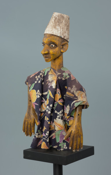 lalka teatralna: Kalhaldi (muzułmański uczony) - Ujęcie z przodu z lewej strony. Lalka teatralna przedstawiająca postać męską w nakryciu głowy w formie walca rozszerzonego u dołu, pomalowanego na biało. Szata z kwiecistego materiału, szeroka, z szerokimi rękawami. Oczy białe, z czarnymi źrenicami, drobne i wypukłe, z zaznaczonymi powiekami dolnymi oraz łukowatymi czarnymi brwiami.  Nos rozszerzony u dołu, usta zamknięte, z dolną wargą pełniejszą. Ręce przy tułowiu. Dłonie z zaznaczonymi palcami.