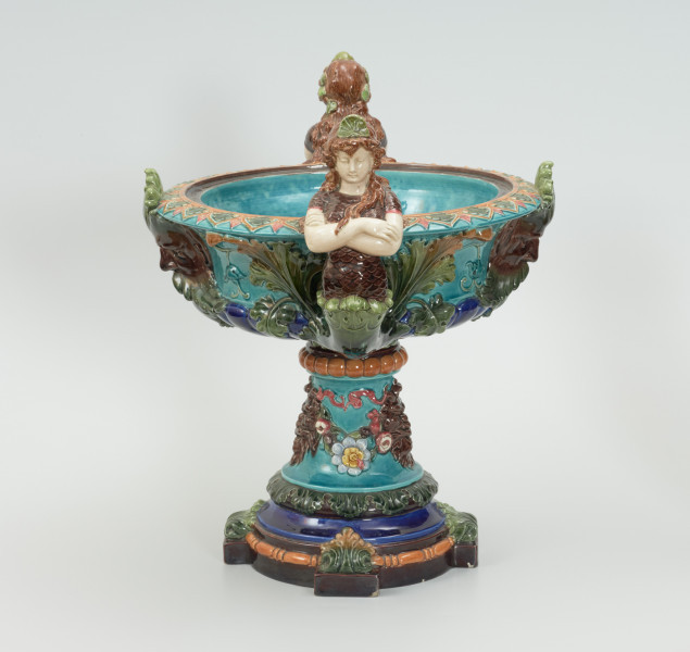 Waza - Ujęcie z prawej strony. Waza w kształcie smukłej amfory. Brzusiec ceramiczny, rozszerzający się ku górze, w kolorze kremowym, malowany podszkliwnie (z uzupełnieniami naszkliwnymi) we wzory kwiatowe - bukiety barwnych kwiatów w typie aster w kolorach fioletowo-czerwonym, liliowym, żółtym i białym, z gałązkami i listkami oraz drobnymi kwiatkami przypominającymi bratki o barwie fioletowo-czerwonej i liliowej. Górna część wazy wraz z uchwytami oraz stopą i część podstawy wykonane jako odlewy z mosiądzu. Mosiężna szyja wąska z otokiem perełkowym w środku ozdobiona jest pionowym, rozszerzającym się ku dołowi i ku górze rytmicznym podziałem z ornamentem geometryczno-roślinnym. Na pogrubionym skraju szyi umieszczona jest płytka miseczka w formie kwiatu o wąskich płatkach, stanowiąca wylewkę wazonu. Poniżej dolnej części szyi oprawa wierzchołka brzuśca składająca się z otoku o sercowym wycięciu zwieńczonym profilowaną palmetką oraz dwóch wydłużonych, sięgających 1/3 wysokości brzuśca, pionowych wsporników uchwytów. Te ostatnie składają się z 2 części: zewnętrzna wychodząca ze wsporników opartych na brzuścu, wyniesiona jest po niewielkim skosie do połowy wysokości szyi i na końcu wywinięta do wnętrza wolutowo, a druga - krótsza, skierowana ku dołowi łączy łagodnym łukiem wolutę z górną częścią oprawy brzuśca, kończąc się wywiniętymi na zewnątrz listkami. Uchwyty żłobkowane, zdobione motywem liści. Wazon wsparty jest na mosiężnej podstawie, składającej się z wieńca oplecionego wstęgą, na którym spoczywa zwężająca się ku górze stopa o brzegach ozdobionych perełkowaniem i pierścieniem, wspierającym koszyczek, ujmujący dolną część ceramicznego brzuśca, w formie kwiatu o ostro zakończonych, wąskich płatkach. Całość spoczywa na podstawie z kwadratowej płytki czerwonego marmuru ze ściętymi narożnikami, ujętymi mosiężnymi wspornikami ramy wspierającej płytę od dołu i opartej w narożach na czterech lwich łapach.