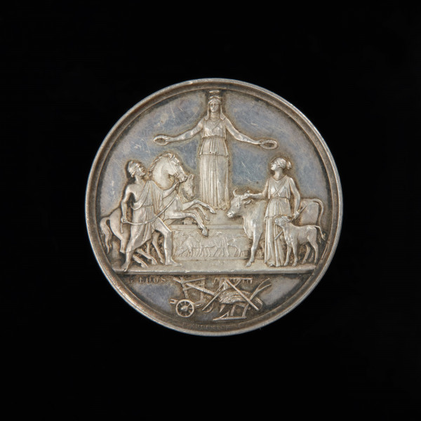 medal nagrodowy; nagroda honorowa - Ujęcie awersu. Na awersie stojąca en face na cokole bogini (Ceres), trzymająca w wyciągniętych rękach dwa wieńce, którymi wieńczy mężczyznę trzymającego wspiętego konia oraz kobietę z krową i cielęciem przy boku. Na cokole wizerunki zwierząt hodowlanych. W dolnej części kompozycja złożona ze snopka i narzędzi rolniczych (pług, grabie, motyka). Z lewej strony kompozycji jedna sygnatura, a wzdłuż rantu druga sygnatura.