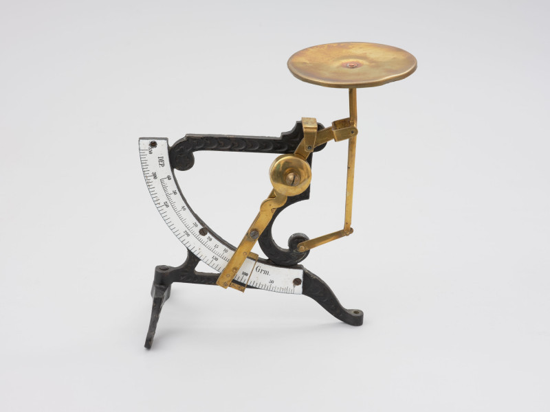 jednowahadłowa uchylna waga pocztowa - Ujęcie z przodu;  Jednowahadłowa uchylna waga pocztowa. Waga składa się ze stalowej skali o wartości od 0 do 350 gram, z podziałką co 5 gram, która połączona jest z trzonem wagi za pomocą stalowych blaszek znitowanych na ruchomo. Podstawa wagi składa się z dwóch elementów, z których element poprzeczny przymocowany jest do głównego elementu na ruchomo, co pozwala składać nóżkę poprzez jej przekręcenie. Dłuższe ramię podstawy zakończone jest śrubką pozwalającą na regulację wagi w opozycji pionowej. Stalowa konstrukcja łącząca główny trzon wagi z dźwignią nośną nasadzona jest na szalkę.