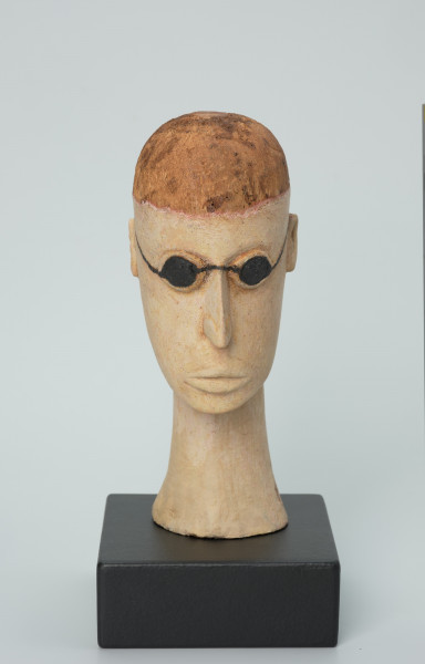 lalka teatralna: Tubabu Cè Kun - Ujęcie z przodu. Drewniane popiersie mężczyzny.  Głowa owalna, wydłużona, zaznaczona linia włosów nad czołem. Włosy pomalowane na kolor jasno-brązowy/blond. Nos długi, wąski, usta wypukle, pomalowane na kolor jasno-różowy. Zamiast oczu czarne okulary, okrągłe, z cienkimi nausznicami. Spod okularów wystające powieki dolne i górne. Szyja długa, wąska, rozszerzona u dołu, pomalowana na kolor jasno-różowy.