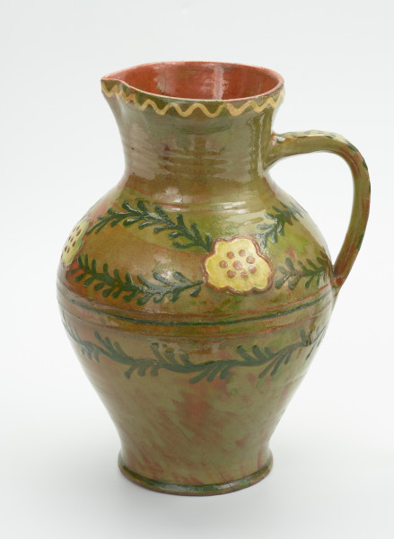 Gliniany dzban zdobiony motywem kwiatowym - Ujęcie z prawej strony. Dzban gliniany. Brzusiec mocno uwypuklony przechodzi w szeroką nieco rozchyloną szyjkę z małym dziobkiem. Ucho taśmowe wzdłużne. Wewnątrz glazura brązowa, na zewnątrz zielona. Dookolny ornament jodełkowy na brzuścu. Krawędź szyjki ozdobiona linią falistą. Ornament wykonany emalią zieloną i żółtą.