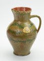 Gliniany dzban zdobiony motywem kwiatowym - Ujęcie z prawej strony. Dzban gliniany. Brzusiec mocno uwypuklony przechodzi w szeroką nieco rozchyloną szyjkę z małym dziobkiem. Ucho taśmowe wzdłużne. Wewnątrz glazura brązowa, na zewnątrz zielona. Dookolny ornament jodełkowy na brzuścu. Krawędź szyjki ozdobiona linią falistą. Ornament wykonany emalią zieloną i żółtą.