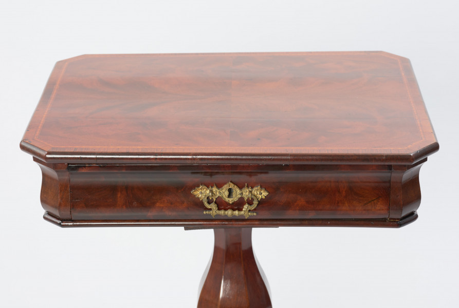 Stolik do robótek - Ujęcie blatu stolika. Stolik z prostokątnym blatem ze ściętymi narożami i sfazowaną krawędzią, ozdobiony intarsją w formie wąskiego otoku z geometrycznym wzorem. Ścianka pod blatem o wybrzuszonej linii przekroju kryje szufladę z mosiężnym okuciem z dziurką do klucza i ruchomym uchwytem w centrum ścianki czołowej. Stolik wsparty na nodze w formie wydłużonej ośmiobocznej tralki gruszkowej, opartej przewężonym profilem na ośmiobocznym trzonie, do którego przymocowane są po przekątnej cztery nóżki składające się z dwóch odwróconych i połączonych esownic. Wewnątrz stolika znajduje się szuflada, która podzielona jest na jedenaście symetrycznie rozmieszczonych przegródek różnej wielkości, z krawędziami podmalowanymi na czarno.