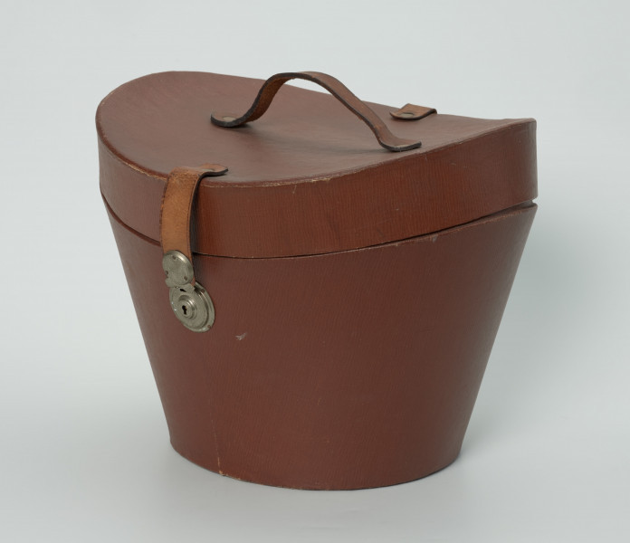 owalny futerał do kapelusza - Ujęcie z z ukosa; Owalny futerał do kapelusza. Owalne, rozszerzające się ku górze pudło z pokrywą o zagłębionej formie z uchwytem w centrum. Pokrywa zamykana na zamek kluczykowy.