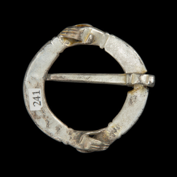 Pozłacana zapinka ze splecionymi dłońmi - Ujęcie z tyłu w poziomie. Pozłacana zapinka, wykonana z dwóch owalnych taśm, w końcach uformowanych w dłonie. Powierzchnia gładka. Kolec wąsk, długi.