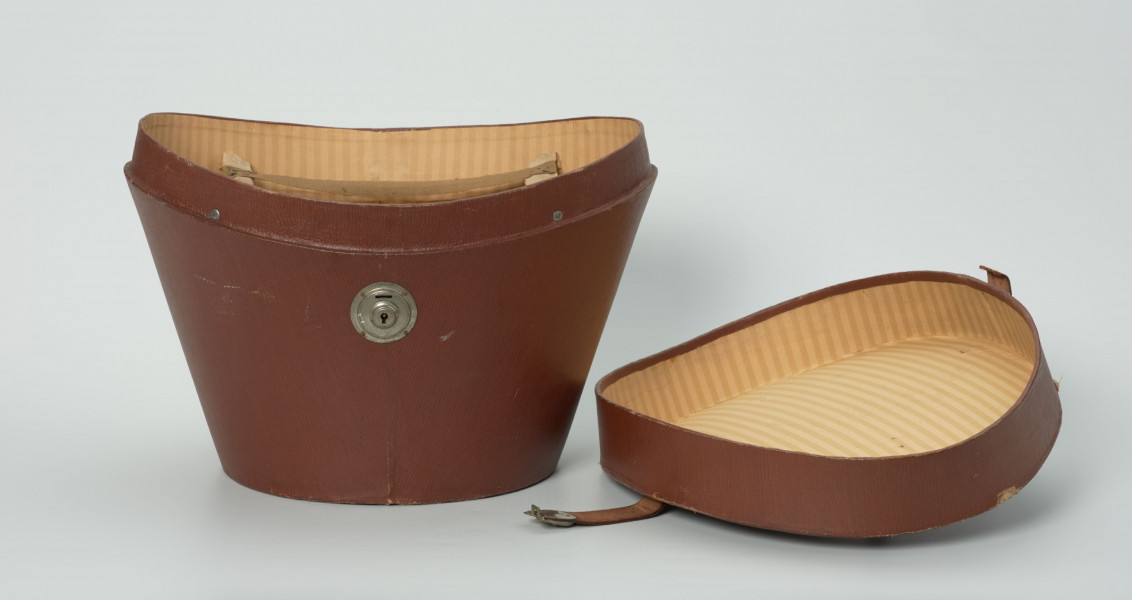 owalny futerał do kapelusza - Ujęcie od przodu z odczepioną pokrywą; Owalny futerał do kapelusza. Owalne, rozszerzające się ku górze pudło z pokrywą o zagłębionej formie z uchwytem w centrum. Pokrywa zamykana na zamek kluczykowy. Wewnątrz podszewka z tkaniny w paski. W górnej części przymocowane pasy z tkaniny do zawieszenia ronda cylindra.