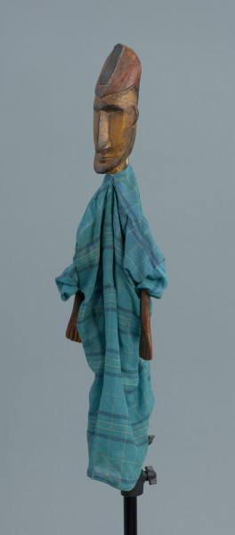 lalka teatralna: Donso (myśliwy) - Ujęcie z przodu skosem w lewą stronę. Drewniana lalka przedstawiająca postać męską. Twarz podłużna, wąska, mocno zarysowane czarne brwi, długi nos, wąskie usta, niewielka broda i wąsy. Postać jest ubrana w długą niebieską szatę w kratę. Na głowie wysoka czapka. Dłonie z zaznaczonymi palcami.