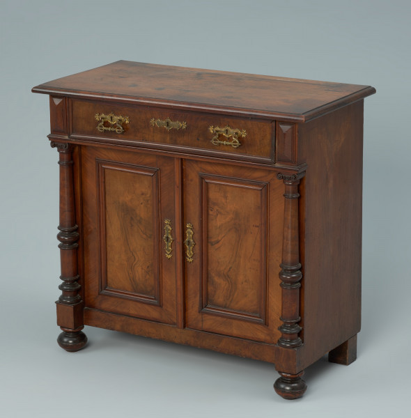 szafka - Ujęcie z przodu, z prawej strony. Szafka w typie bieliźniarki z drewna fornirowanego, przeznaczona pod lustro, dwudrzwiowa z szufladą górną. Drzwi bieliźniarki ujęte o bokach dwiema toczonymi i profilowanymi kolumnami z kapitelami w typie jońskim o żłobkowanych poprzecznie wolutach. Kolumienki wsparte są na pionowych kostkach, wysuniętych przed lico skrzyni dolnej, pod którymi znajdują się toczone kuliste nogi z profilem w górnej części. Ponad kolumnami na gzymsie kryjącym dno szuflady umieszczono flankujące ją pionowe kaboszony. Czoło szuflady stanowiącej podstawę blatu szafki, ujęte na krawędziach profilowaną listwą. Blat z uskokiem i półkolistym sfazowaniem na brzegu. Z tyłu blatu dwa otwory do kołków trzymających lustro. Drzwi z profilowaną listwą przymykową posiadają w środku zagłębioną, prostokątną płycinę wydzieloną profilowaną listwą, okalającą zewnętrzną ramę. Wydłużone okucia mosiężne, złocone z otworem do klucza w układzie pionowym na obu skrzydłach drzwi. Trzy analogiczne okucia umieszczone poziomo na szufladzie - w centrum z otworem do klucza, a po bokach z ruchomym uchwytem, zdobionym motywem woluty z głową smoka. Tylne nogi proste, klockowe.