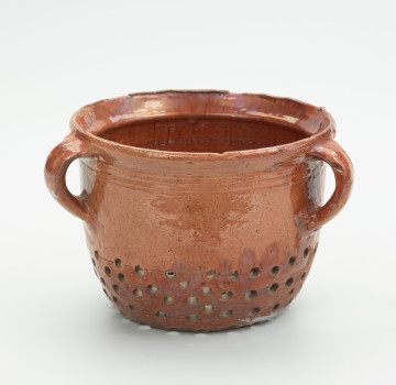 Gliniany durszlak z trzema uchami - Ujęcie z przodu. Naczynie kuchenne z gliny. Ścianki proste, przy dnie lekko zaokrąglone. Krawędź wylewu nieznacznie wygięta na zewnątrz. Trzy ucha taśmowate, mocowane do krawędzi wylewu w połowie wysokości naczynia. Od dołu 1/3 powierzchni durszlaka dziurkowana. Pod krawędzią wylewu naczynie zdobione dookolnym rytem. Cztery dookolne ryty znajdują się również w dziurkowanej części.