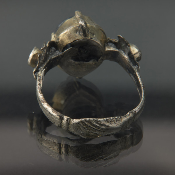 Pozłacany pierścień z niebieskim oczkiem - Ujęcie z tyłu. Srebrny pierścień. Z przodu splecione dłonie w długich rękawach. Z tyłu, na środku srebrny owal z kołnierzem, po bokach dwa srebrne okręgi ze srebrnymi płatkami.