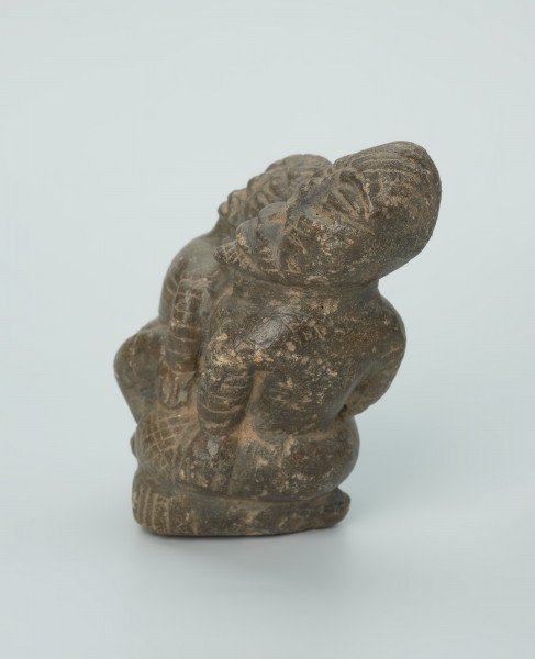 figura kultu przodków - Ujęcie z boku z lewej. Rzeźbione w grafitowym kamieniu dwie postaci ludzkie w pozycji siedzącej. Mniejsza postać – najprawdopodobniej dziecko - siedzi na kolanach większej - najprawdopodobniej kobiety. Obie postacie ozdobiono licznymi nacięciami liniowymi. Widoczne rysy i mikropęknięcia.