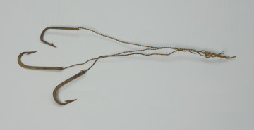 haczyki - 3 szt. - Ujęcie przodu haczyków z góry. Haczyk (3 szt.). Wyrób fabryczny, wykonany z mosiądzu.  Trzonek haczyka prosty, spłaszczony, bez dodatkowych zadziorów z niewielkim oczkiem, w którym mocowany jest wykonany ze skręconego drutu, przypon. Grot prosty (znajdujący się w jednej płaszczyźnie z trzonkiem), ze znajdującym się po jego wewnętrznej stronie zadziorem.