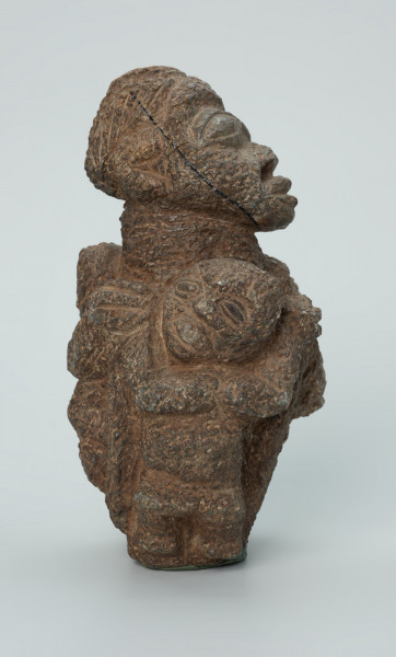 rzeźba; przedmiot obrzędowy; figura kultu zmarłych - Ujęcie prawy bok. Rzeźbiona w ziemistym steatycie siedząca postać ludzka o cechach kobiecych, otoczona przez trójkę mniejszych, dzieciecych postaci, skierowanych ku niej z uniesionymi rękoma.