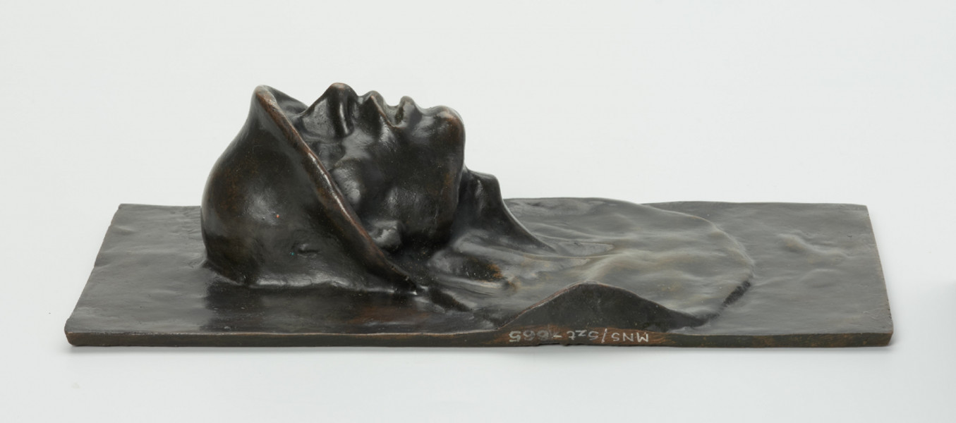 portret męski - Ujęcie w poziomie z prawej strony; Płaskorzeźba przedstawiająca głowę mężczyzny. Z blachy stanowiącej tło wyłania się kwadratowa twarz z szerokim nosem i rozchylonymi ustami, szyja mocno osadzona, szeroka. Na głowie hutniczy kask. Widoczny fragment torsu.