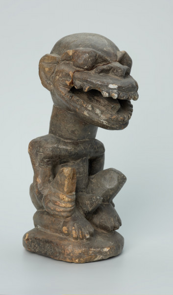 rzeźba; przedmiot obrzędowy; Figura kultu zmarłych - Ujęcie z przodu z prawej strony. Rzeźbiona w grafitowym steatycie, usadowiona na płaskiej, owalnej podstawie postać siedzącego z podkurczonymi nogami mężczyzny o masywnej głowie i tęgiej szyi z odsłoniętymi genitaliami. Powierzchnia rzeźby niejednolita; ubytek na prawym kolanie; ślady klejenia na twarzy.