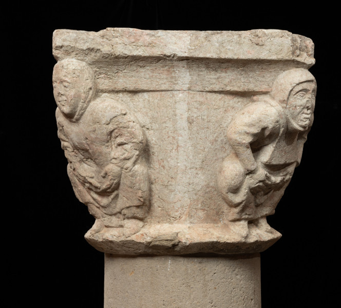 Kapitel z przedstawieniem czterech mnichów - atlantów - Ujęcie jednego boku skosem. Kapitel blokowo-kielichowy, rozszerzający się ku górze, wykuty z jednego kawałka kamienia. Okrągłą podstawę otacza wałek o trójkątnym profilu. Zwieńczenie w formie szerokiej czworobocznej płyty abakusa. W narożnikach kapitela płaskorzeźbione wizerunki czterech mnichów. Postaci o krępej budowie ukazane frontalnie, w pozycji przykucniętej. Stopami opierają się o dolny wałek, głowami podpierają abakus. Relief głęboki, półplastyczny, światłocieniowy. Gładko opracowane tło. Niewielkie uszczerbienia zwłaszcza w partii abakusa i dolnego wałka. Pod spodem centralne wgłębienie do umocowania trzpienia łączącego kapitel z trzonem.