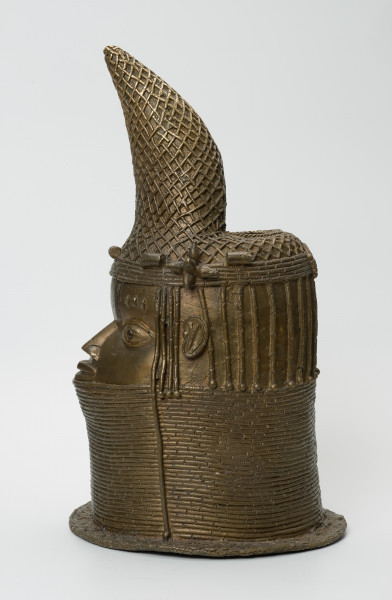 Głowa Królowej Matki - Ujęcie z boku z lewej strony. Brązowy, pusty w środku odlew głowy kobiety, o złotym zabarwieniu. Twarz o cechach negroidalnych, wydatnych, lekko rozchylonych ustach oraz szerokim u nasady nosie ma oczy z wyraźnie zaznaczoną linią powiek. Na ich wysokości usytuowane są nieduże uszy. Lekko wypukłe czoło dekorowane jest pionowymi inkrustacjami z żelaza oraz trzema podłużnymi, symetrycznie rozmieszczonymi, pogrubieniami nad każdym okiem. Szyję i podbródek do wysokości dolnej wargi zasłonięto masywnym naszyjnikiem z korali. Głowa jest nienaturalnie poszerzona w tylnej części, tuż pod przechyloną do przodu koroną o charakterystycznym kształcie dziobu papugi, którą nad uszami ozdobiono dwoma rozetami. W dolnej części korony widnieje pięć ozdób o walcowatej formie, trzy nad czołem, dwie za rozetami oraz opaska z korali. Tuż za koroną znajduje się duży, owalny otwór na rzeźbiony kieł słonia, którego brak. Poniżej, po obu stronach twarzy i z tyłu głowy, zaznaczono zaplecione w warkoczyki włosy. Z tyłu głowy pomiędzy włosami a naszyjnikiem usytuowany jest niewielki, okrągły otwór. U podstawy odlewu znajduje się pozioma listwa, dekorowana motywem plecionki.