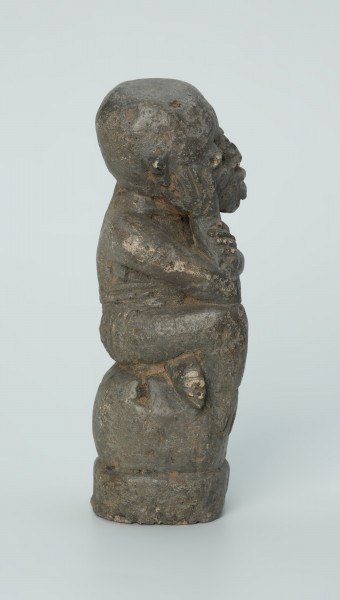 figura kultu przodków - Ujęcie z boku z prawej. Rzeźbiona w grafitowym kamieniu postać ludzka w pozycji siedzącej - najprawdopodobniej mężczyzny. Postać siedzi na wysokiej podstawie, która swym kształtem przypomina kamień. Rzeźba ma charakterystyczne poziome nacięcia widoczne na obu policzkach. Widoczne rysy, mikropęknięcia i liczne malutkie otworki.