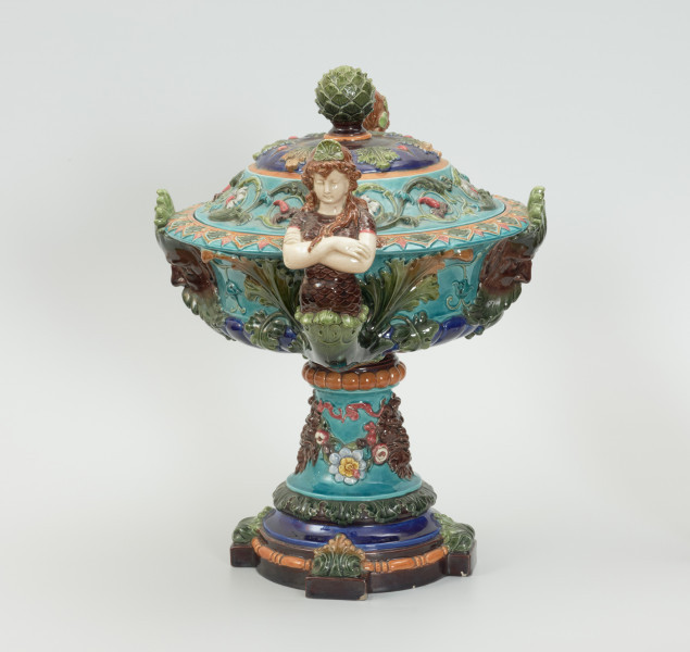 Waza - Ujęcie z prawej strony z pokrywką. Waza w kształcie smukłej amfory. Brzusiec ceramiczny, rozszerzający się ku górze, w kolorze kremowym, malowany podszkliwnie (z uzupełnieniami naszkliwnymi) we wzory kwiatowe - bukiety barwnych kwiatów w typie aster w kolorach fioletowo-czerwonym, liliowym, żółtym i białym, z gałązkami i listkami oraz drobnymi kwiatkami przypominającymi bratki o barwie fioletowo-czerwonej i liliowej. Górna część wazy wraz z uchwytami oraz stopą i część podstawy wykonane jako odlewy z mosiądzu. Mosiężna szyja wąska z otokiem perełkowym w środku ozdobiona jest pionowym, rozszerzającym się ku dołowi i ku górze rytmicznym podziałem z ornamentem geometryczno-roślinnym. Na pogrubionym skraju szyi umieszczona jest płytka miseczka w formie kwiatu o wąskich płatkach, stanowiąca wylewkę wazonu. Poniżej dolnej części szyi oprawa wierzchołka brzuśca składająca się z otoku o sercowym wycięciu zwieńczonym profilowaną palmetką oraz dwóch wydłużonych, sięgających 1/3 wysokości brzuśca, pionowych wsporników uchwytów. Te ostatnie składają się z 2 części: zewnętrzna wychodząca ze wsporników opartych na brzuścu, wyniesiona jest po niewielkim skosie do połowy wysokości szyi i na końcu wywinięta do wnętrza wolutowo, a druga - krótsza, skierowana ku dołowi łączy łagodnym łukiem wolutę z górną częścią oprawy brzuśca, kończąc się wywiniętymi na zewnątrz listkami. Uchwyty żłobkowane, zdobione motywem liści. Wazon wsparty jest na mosiężnej podstawie, składającej się z wieńca oplecionego wstęgą, na którym spoczywa zwężająca się ku górze stopa o brzegach ozdobionych perełkowaniem i pierścieniem, wspierającym koszyczek, ujmujący dolną część ceramicznego brzuśca, w formie kwiatu o ostro zakończonych, wąskich płatkach. Całość spoczywa na podstawie z kwadratowej płytki czerwonego marmuru ze ściętymi narożnikami, ujętymi mosiężnymi wspornikami ramy wspierającej płytę od dołu i opartej w narożach na czterech lwich łapach.