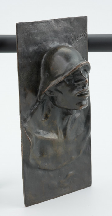 portret męski - Ujęcie z przodu skosem w prawą stronę; Płaskorzeźba przedstawiająca głowę mężczyzny. Z blachy stanowiącej tło wyłania się kwadratowa twarz z szerokim nosem i rozchylonymi ustami, szyja mocno osadzona, szeroka. Na głowie hutniczy kask. Widoczny fragment torsu.