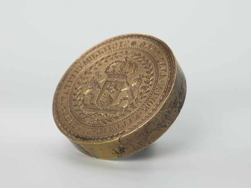 tłok pieczętny - Ujęcie z przodu skosem w lewą stronę. Okragły tłok pieczętny z brązu z trzpieniem montarzowym oraz ukoronowaną tarczą herbową Szczecina trzymaną przez dwa lwy w polu pieczęci.