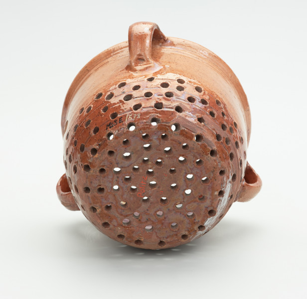 Gliniany durszlak z trzema uchami - Ujęcie z tyłu w poziomie dnem do przodu. Gliniane, zaokrąglone naczynie, z dziurkami rozmieszczonymi nieregularnie na zaokrąglonym tyle. Na zewnątrz trzy uszka. Na powierzchni widoczny połysk.