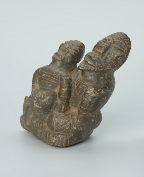 figura kultu przodków - Ujęcie z przodu z lewej skosem. Rzeźbione w grafitowym kamieniu dwie postaci ludzkie w pozycji siedzącej. Mniejsza postać – najprawdopodobniej dziecko - siedzi na kolanach większej - najprawdopodobniej kobiety. Obie postacie ozdobiono licznymi nacięciami liniowymi. Widoczne rysy i mikropęknięcia.