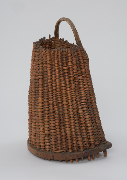 Kosz do przenoszenia ryb - Ujęcie z przodu skosem w lewą stronę. Kosz do przenoszenia ryb o kształcie ściętego stożka z tylną ścianką prostą. Wykonany z dartych korzeni sosny techniką żeberkowo-krzyżową – sztywnych szkielet z tzw. żeberek jest gęsto opleciony giętkimi wiciami „osnowy”. Podstawa kosza wycięta z jednego kawałka drewna o grubości ok. 2 cm, półokrągła. Wzdłuż jej krawędzi biegnie szereg otworów, w które wchodzą żeberka. Ich wystające końce tworzą rodzaj krótkich nóżek. Dłuższe krawędzie tylnej ścianki stanowi wygięta w pałąk, wystająca poza górną krawędź kosza okorowana gałąź (uchwyt). Mocowana jest do podstawy za pomocą gwoździ.