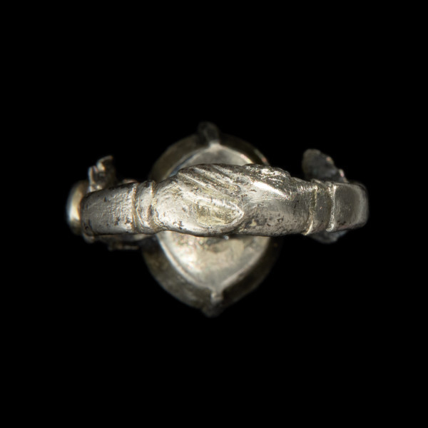 Pozłacany pierścień z niebieskim oczkiem - Ujęcie tyłu w powiększeniu. Srebrny okręg ze splecionymi dłońmi w długich rękawach. U dołu płaski owal z kołnierzem.