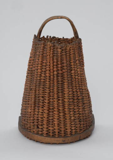 Kosz do przenoszenia ryb - Ujęcie z przodu. Kosz do przenoszenia ryb o kształcie ściętego stożka z tylną ścianką prostą. Wykonany z dartych korzeni sosny techniką żeberkowo-krzyżową – sztywnych szkielet z tzw. żeberek jest gęsto opleciony giętkimi wiciami „osnowy”. Podstawa kosza wycięta z jednego kawałka drewna o grubości ok. 2 cm, półokrągła. Wzdłuż jej krawędzi biegnie szereg otworów, w które wchodzą żeberka. Ich wystające końce tworzą rodzaj krótkich nóżek. Dłuższe krawędzie tylnej ścianki stanowi wygięta w pałąk, wystająca poza górną krawędź kosza okorowana gałąź (uchwyt). Mocowana jest do podstawy za pomocą gwoździ.