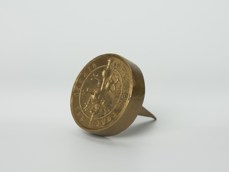 tłok pieczętny - Ujęcie z przodu skosem w lewą stronę rysunkiem gryfa do przodu w dół. Okrągły tłok pieczętny z brązu z trzpieniem montarzowym w formie ostrosłupa oraz rysunkiem gryfa na iglicy wieży w polu pieczęci.