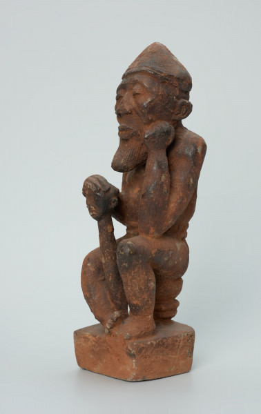 Ujęcie z przodu z lewej. Rzeźbiona w brunatno-czerwonym kamieniu dość duża postać mężczyzny (starca?) w pozycji siedzącej. Mężczyzna siedzi na okrągłym, malutkim stołku, ma charakterystyczne nakrycie głowy - spiczastą czapkę o zaokrąglonych rogach. Jego prawa ręka opiera się o berło (laskę) zakończone rzeźbą głowy. Widoczne rysy, mikropęknięcia i liczne malutkie otworki.