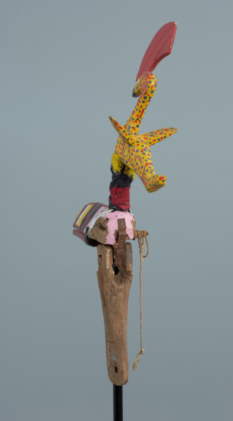 Ujęcie z tyłu. Kolorowy ptak zrobiony z drewna z ruchomą żuchwą poruszaną za pomocą sznurka. Długi, pomalowany w kolorowe paski dziób. Na głowie umieszczony drugi, mniejszy ptak w żółtym kolorze z czerwonym czubem.